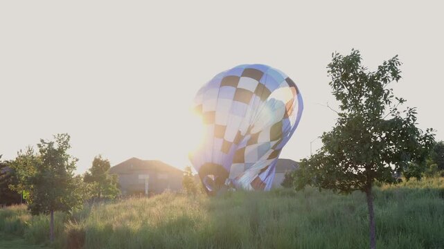 Large blue and brown checkered hot air balloon touching down and deflating in a grassy field near a suburban neighborhood at sunset