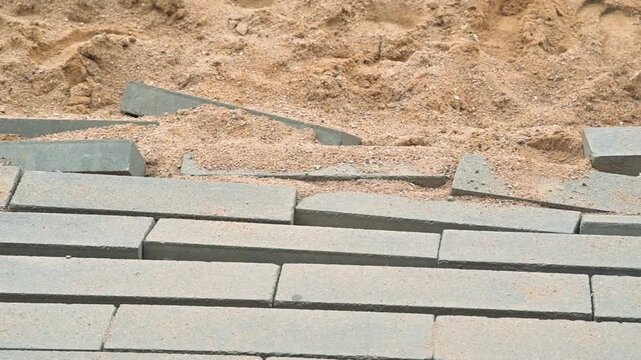 gray paving stones on a sandy surface to create a pathway. The action involves arranging the stones in a systematic manner over the sand