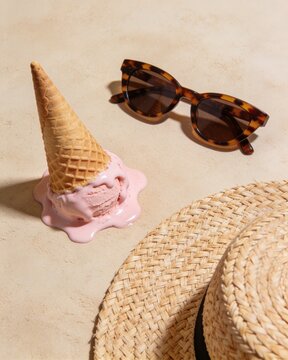 High angle flat lay of an upside-down melting strawberry ice cream cone, tortoiseshell sunglasses, and a woven straw hat on warm sand during summer vacation.