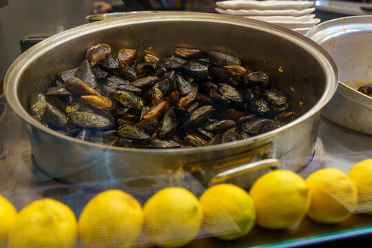 cooked mussels in a pot on a kitchen counter with lemons and plates nearby, close-up view of food preparation in a restaurant kitchen setting, brown and yellow tones, cooking mood, detailed.