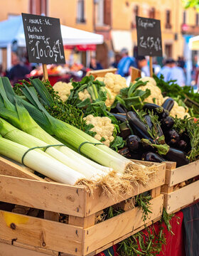 Fresh organic vegetables, including leeks, cauliflower, and eggplants, are displayed in rustic wooden crates at a vibrant European street market, symbolizing healthy, sustainable living.