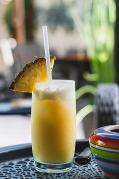 A close-up shot of a tropical yellow cocktail, such as a pina colada or fresh pineapple smoothie, served in a tall glass with a straw and garnished with a pineapple wedge. The drink sits on a black te