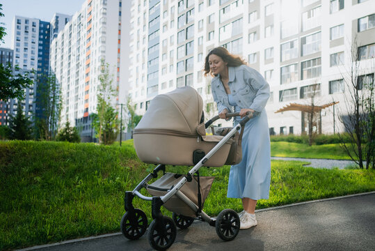 Young mother pushing a baby stroller on a path, looking down at her child with affection, while walking through a contemporary urban park surrounded by modern apartment buildings
