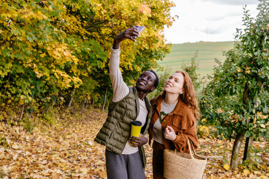 Cheerful multiracial female friends taking smartphone selfie together in colorful autumn orchard