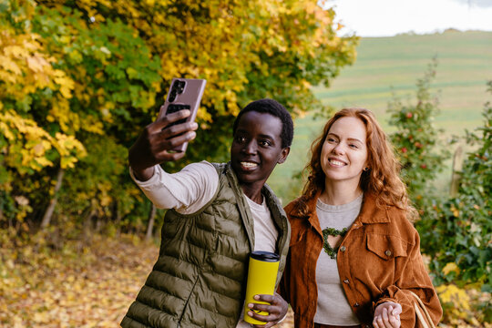 Smiling diverse female friends making smartphone selfie during relaxed autumn orchard walk