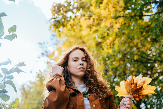 Confident freckled red-haired woman holding colorful leaves in sunny autumn orchard portrait