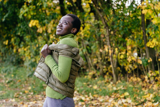 Hopeful Black woman adjusting warm vest during peaceful autumn walk with natural copy space