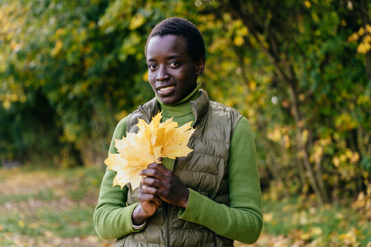 Cheerful Black woman holding yellow autumn leaves during peaceful seasonal walk in nature