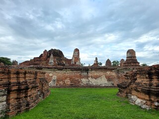 Fototapeta premium Ancient Ruins of Ayutthaya: A breathtaking panorama of ancient ruins in Ayutthaya, Thailand, featuring weathered brick structures, crumbling stupas, and lush green grass under a dramatic cloudy sky.