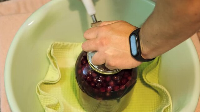 A man canning cherry compote using a manual seaming machine to seal a large glass jar with metal lid. Home canning, food preservation, traditional kitchen, winter storage preparations. A Can Seamer.