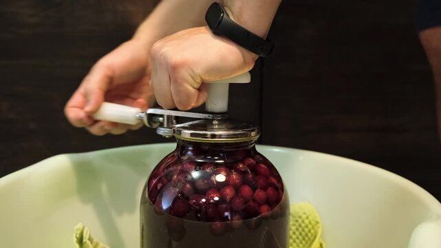A man canning cherry compote using a manual seaming machine to seal a large glass jar with metal lid. Home canning, food preservation, traditional kitchen, winter storage preparations. A Can Seamer.
