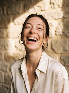 Portrait of a Joyful Brunette Woman Laughing Heartily while Leaning Against a Sunlit Textured Stone Wall
