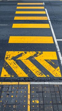 A close-up view of a road with faded yellow pedestrian crossing markings and arrows on a wet asphalt surface