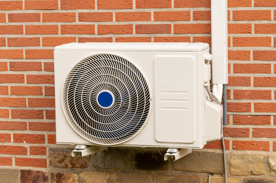 Modern outdoor air conditioner unit installed on a red brick house wall. High-efficiency split system heat pump, residential climate control technology, and smart home ventilation concept