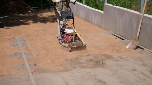 Worker guides plate compactor across dusty soil beside smooth retaining wall and metal railings, with shovel, wheelbarrow, marked guide lines, and soil piles visible in bright