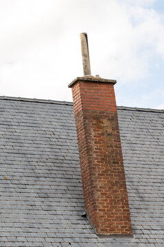A vertical medium shot of a traditional red brick chimney structure rising from a weathered grey slate tile roof. The chimney features a concrete cap and a single tall pipe extension against a cloudy 