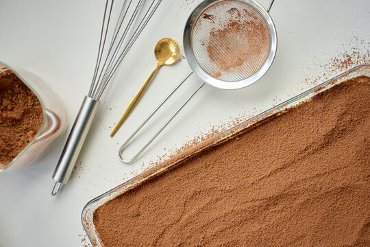 Tiramisu dessert covered with cocoa powder in baking dish, with sieve and cocoa bowl