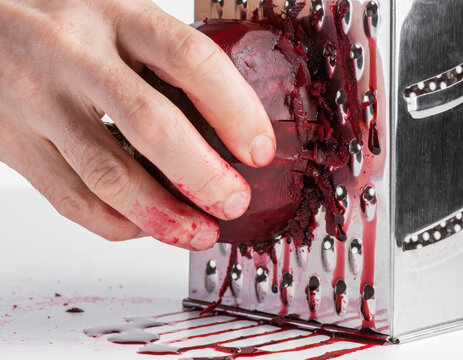 Close up view of a human hand vigorously grating a large, raw red beetroot onto a kitchen grater, resulting in intense ruby red juice and pulp spreading across the white surface
