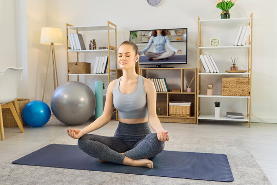 Woman meditation yoga at home with television. She sits cross legged on a mat, eyes shut, following an online class, encouraging mindfulness and balance. Clear concept of mindful home fitness