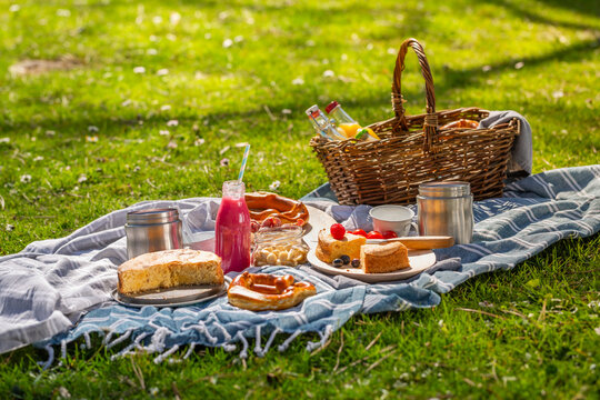 A woven basket filled with beverages and treats sits on a checkered blanket laid out on lush green grass