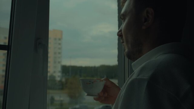 A man drinks tea and looks out the window, where he sees cloudy weather. Tea on a sad day.