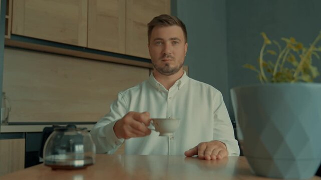 A man drinks tea or coffee with a serious face in the living room and looks attentively at the camera. Video for illustrating home time, a handsome man, English style, VIP persona.