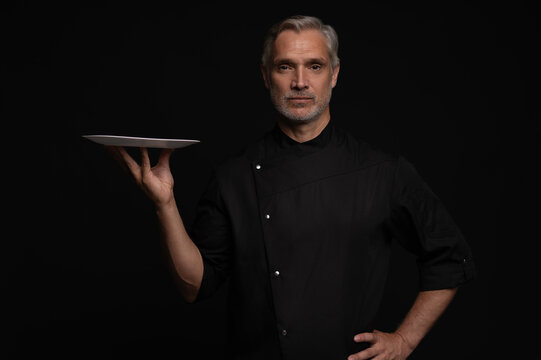 Portrait Of 50s Mature Chef In Uniform Holding Empty Plate on black.