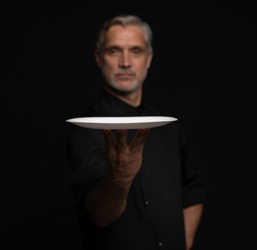 Portrait Of 50s Mature Chef In Uniform Holding Empty Plate on black.