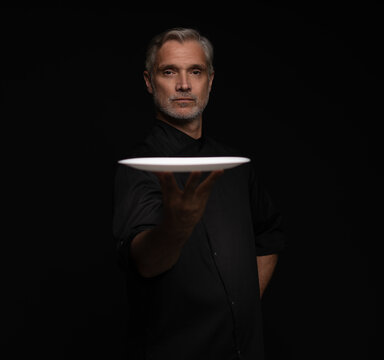Portrait Of 50s Mature Chef In Uniform Holding Empty Plate on black.