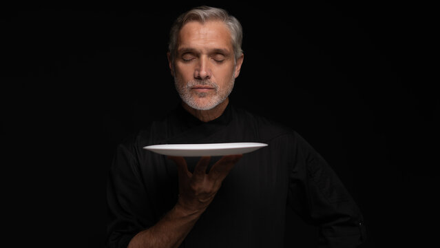 Portrait Of 50s Mature Chef In Uniform Holding Empty Plate on black.
