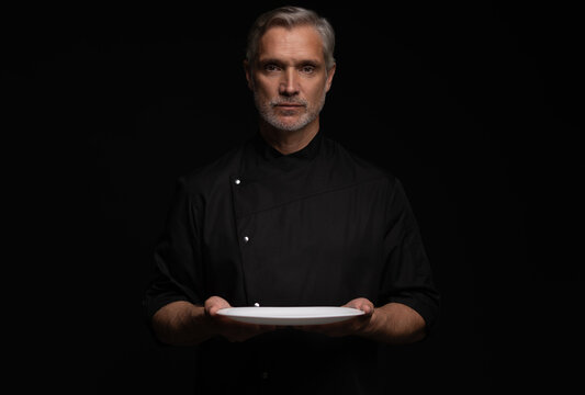 Portrait Of 50s Mature Chef In Uniform Holding Empty Plate on black.