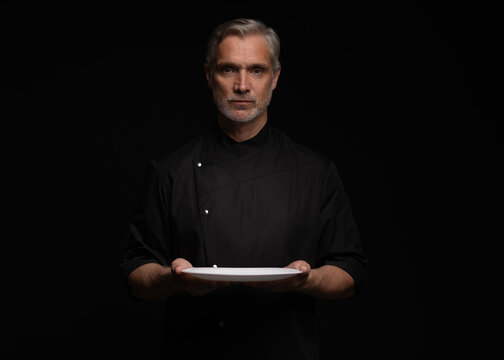 Portrait Of 50s Mature Chef In Uniform Holding Empty Plate on black.