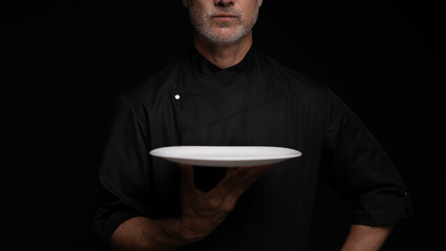 Portrait Of 50s Mature Chef In Uniform Holding Empty Plate on black.