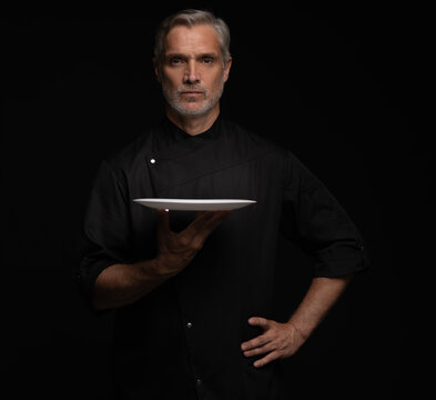 Portrait Of 50s Mature Chef In Uniform Holding Empty Plate on black.