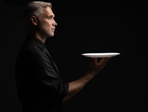 Portrait Of 50s Mature Chef In Uniform Holding Empty Plate on black.