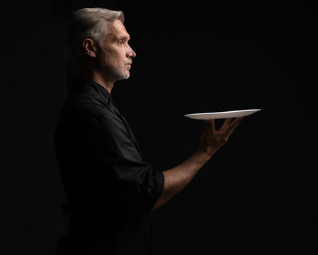 Portrait Of 50s Mature Chef In Uniform Holding Empty Plate on black.