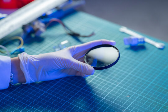 A person in a lab coat holds a lens while standing on a green work surface filled with tools and devices