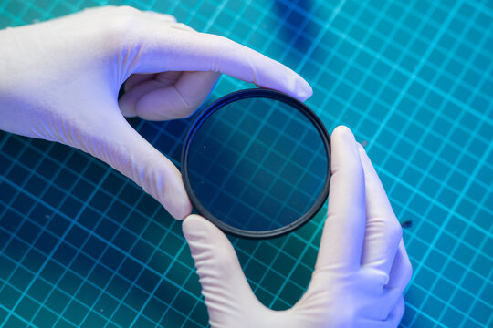 A person is holding a clear lens with gloved hands in a laboratory setting on a green cutting mat