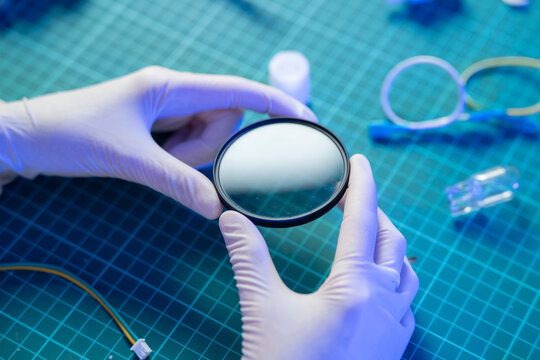 A person wearing gloves examines a round optical lens in a laboratory. Several tools and items are nearby