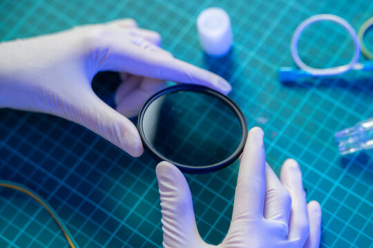 Hands in gloves hold a round lens while working on optics in a laboratory setting with various tools around