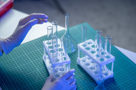 A person wearing gloves is arranging glass test tubes and flasks on a green workspace for vaccine research tasks