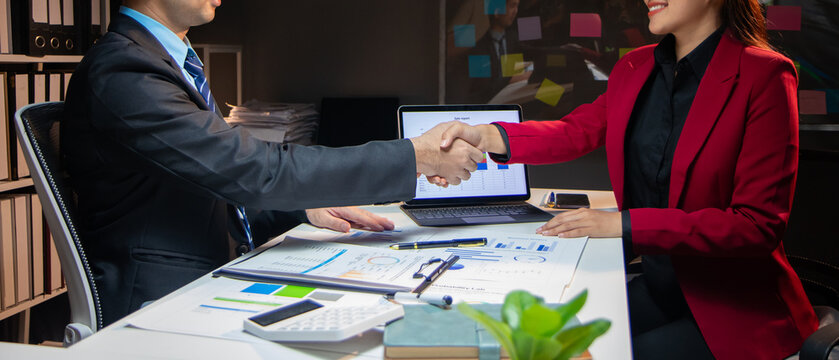 Two business people shaking hands after signing a contract at office desk with laptop, documents, calculator, and financial charts, celebrating successful partnership.