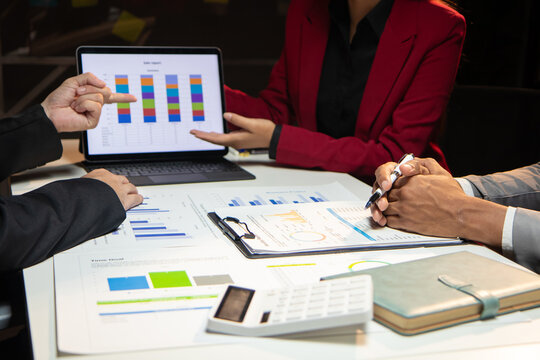Asian businesswoman showing sales data on a laptop screen, presenting reports to colleagues during a late-night meeting in a modern office, teamwork and data analysis.