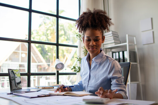 Young African American businesswoman using calculator and taking notes while working on financial report in modern office with laptop and documents on desk.
