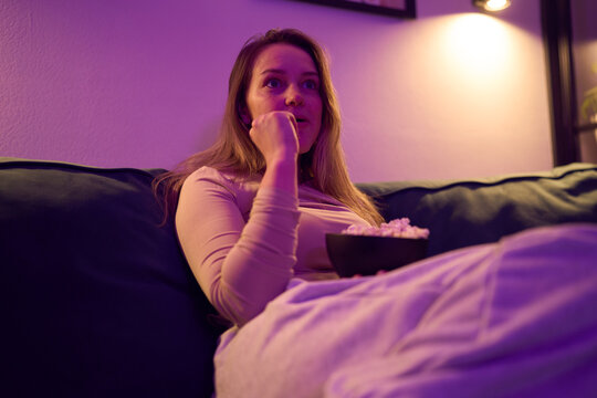 Female watching screen on sofa holding popcorn in evening light