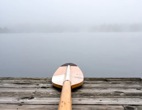 Wooden canoe paddle resting on a weathered wooden dock overlooking a calm, foggy lake. Equipment for outdoor recreation, water sports, and lake activities.