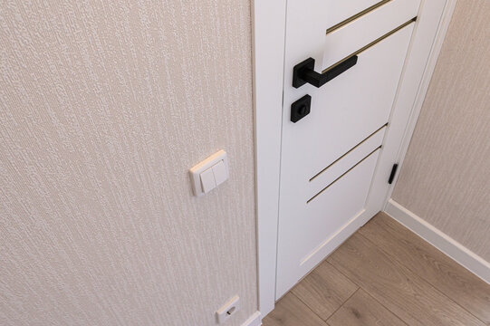 white interior door with minimalist paneling features sleek black lever handle and matching lock. adjacent beige textured walls hold light switch, while light‑wood flooring completes clean entryway