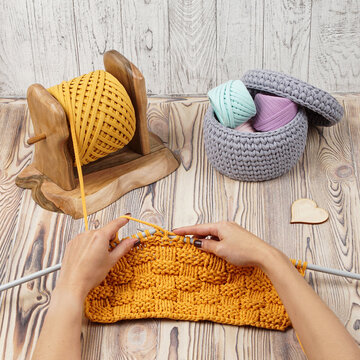  Hands knitting with thick cotton yarn using white needles on a wooden surface. A wooden yarn holder and crocheted basket with colorful spools create a cozy handmade craft composition.