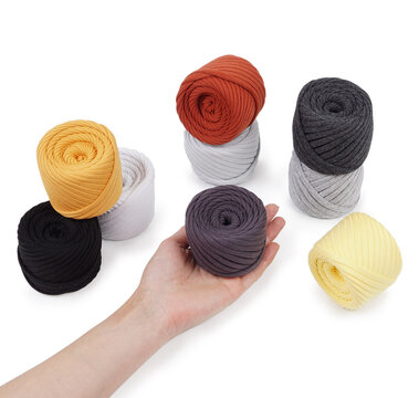 Product photography showing a hand holding a yellow skein of soft thick cotton yarn. Assorted neutral and bright yarn balls on white background.