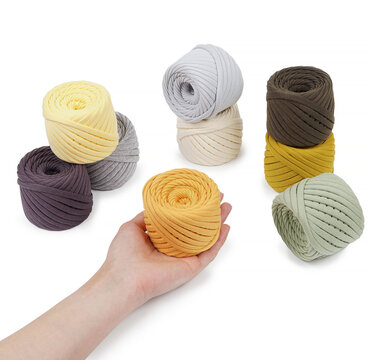   Human hand holding a bright yellow skein of thick cotton t-shirt yarn. Various neutral and pastel yarn balls arranged on white background.
